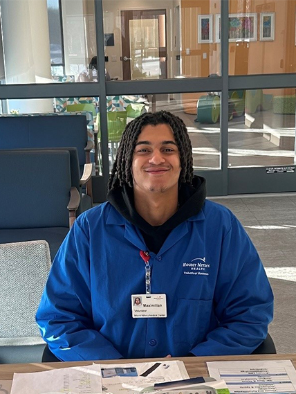 Mount Nittany Health Volunteer, Maximilian Croke sitting behind welcome desk, smiling